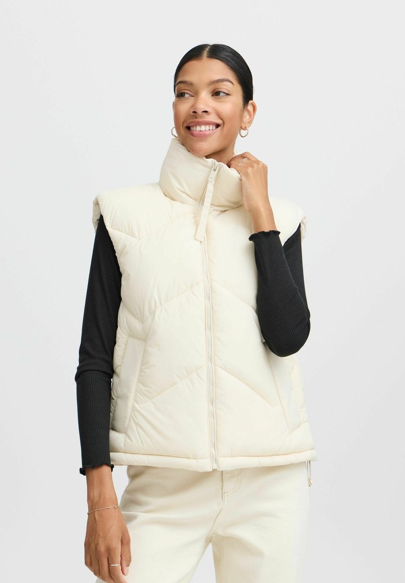 Crème puffer vest met hoge kraag, gevoerd patroon en ritssluiting aan de voorkant. Draag je over een zwarte ribgebreide langemouwen top.