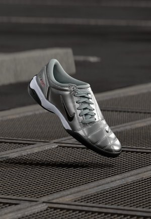 Chaussure de sport argentée avec semelle noire et design à lacets, suspendue au-dessus d'une grille métallique.