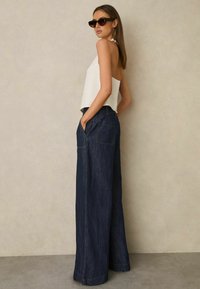 Bezrekawnik w białym kolorze o zwiewnym kroju, zestawiony z szerokimi, ciemnymi spodniami dżinsowymi z dużymi kieszeniami i kontrastowymi przeszyciami.