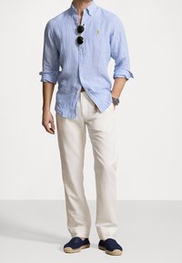 Světle modrá pruhovaná košile na knoflíky, bílé lněné kalhoty, tmavě modré espadrilky a sluneční brýle. Košile má límec a ohrnuté rukávy.