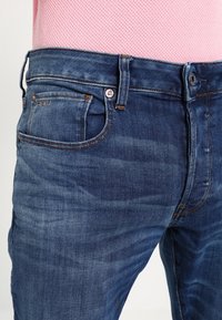 Mörkblå denimjeans med smal passform, med en klassisk femfickdesign, orange sömmar och synlig whiskering på tyget.