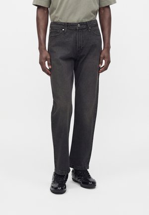 Jean noir droit porté par une personne, associé à une chemise verte et des baskets noires brillantes sur un fond uni.