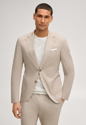 Man met een beige pak, een wit pochet en een licht gestructureerde trui, staande met één hand in de zak tegen een effen achtergrond.