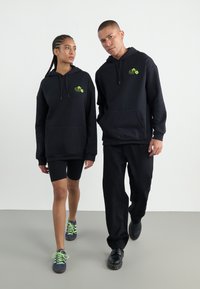 Black hoodies with front pockets and green logos, paired with black cycling shorts and contrasting sneakers, displayed by two figures walking.