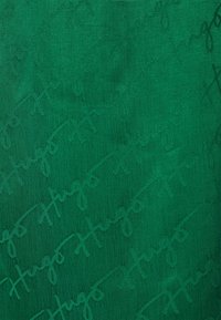 Tela verde con una textura suave que presenta un patrón tonal repetitivo de la palabra "Hugo" en una elegante tipografía.