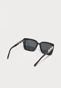 Óculos de sol quadrados pretos com lentes escuras. Apresentam uma haste texturizada com três pedras preciosas transparentes decorativas. Acabamento liso e brilhante nas hastes.