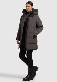 Woman stands wearing a long, dark gray padded coat with a hood, black leggings, and black chunky boots against a plain light background.