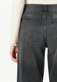 Nærbilde av en persons nedre rygg og hofter iført høyhalsede, mørkegrå denimjeans med baklommer og en lys beige langermet topp.