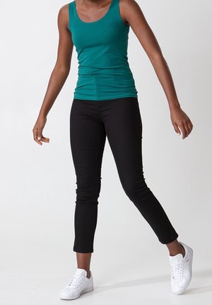 Persoon die een groene mouwloze top, zwarte strakke broek en witte sneakers draagt, staand met één been licht opgetild tegen een effen achtergrond.