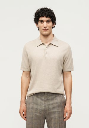 Mand med krøllet hår iført beige kortærmet polo-shirt og grå ternede bukser, står foran en ensfarvet lys baggrund.