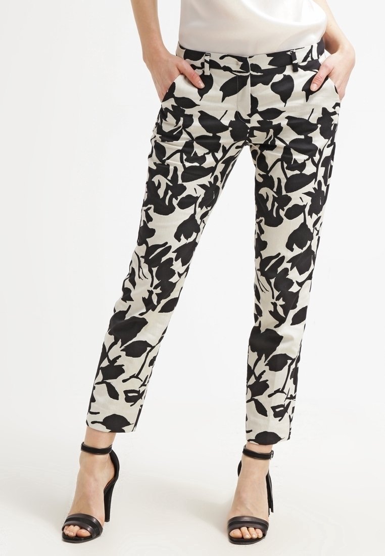 Femme portant un pantalon noir et blanc à motif floral, les mains dans les poches et des sandales noires à talons hauts, debout sur un fond blanc.