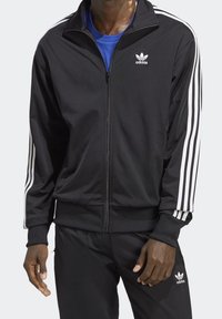 Man i svart Adidas träningsjacka och byxor med vita ränder på ärmarna och Adidas-logotyper, som står framför en neutral bakgrund.