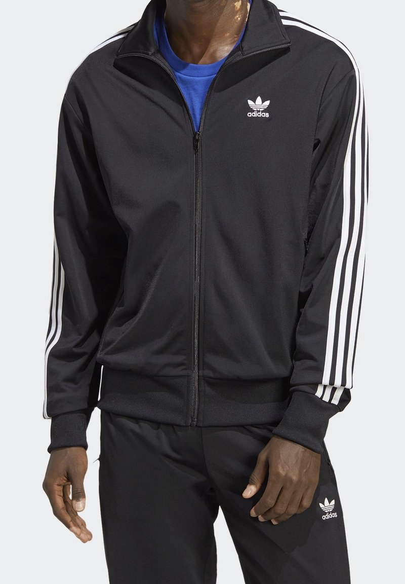 Man i svart Adidas träningsjacka och byxor med vita ränder på ärmarna och Adidas-logotyper, som står framför en neutral bakgrund.