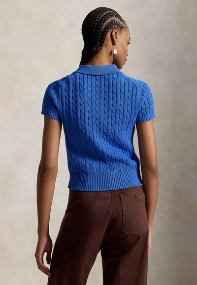 Femme aux cheveux tressés portant un haut bleu à manches courtes en maille torsadée et un pantalon marron, debout de dos face à la caméra sur un fond neutre.