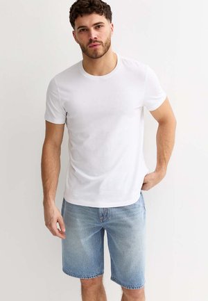 Homme aux cheveux courts et bouclés avec une barbe, portant un t-shirt blanc et un short en denim bleu clair, debout avec une main dans la poche.