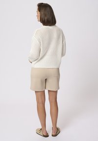 Pull en maille blanche léger avec tricot ajouré, associé à un short beige. Ourlet de shorts à revers visible et des chaussures plates à enfiler.