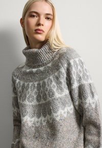 Pull en tricot gris avec un col haut, présentant un motif géométrique blanc et des fils multicolores à travers le tissu.