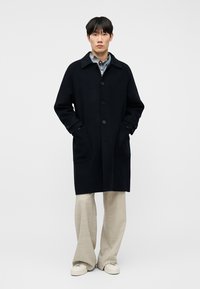 Άνδρας με μακρύ σκούρο μπλε παλτό πάνω από μπλε πουκάμισο με καρό, μπεζ παντελόνι φαρδιάς γραμμής και λευκά αθλητικά παπούτσια, στέκεται μπροστά σε απλό λευκό φόντο.