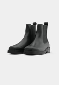 Melnas ādas Čelsijas zābaki ar elastīgām sānu daļām, noapaļotiem pirkstiem un vilkšanas cilpiņām pie papēža; plakani gumijas zoles ar minimālu protektoru.