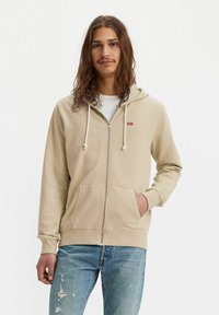 Junger Mann mit langen Haaren trägt einen beigen Reißverschluss-Hoodie mit Levi's-Logo und zerrissene blaue Jeans, steht vor einem weißen Hintergrund.