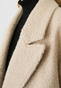 Beige textured wool coat with a notched collar, shown alongside a black ribbed turtleneck, highlighting soft material and warm design.