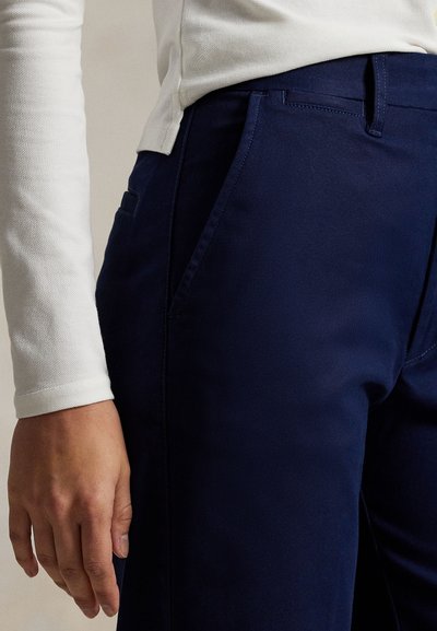 Person som har på seg marineblå bukser og en hvit langermet topp, med venstre hånd avslappet ved siden av.