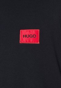 Czarny sweter z dzianiny z czerwoną prostokątną metką z logo „HUGO” w czarnym tekście, umieszczoną w górnym lewym rogu.