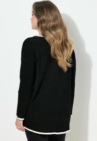 Černý svetr s volnějším střihem, s krémově zbarvenými lemy na manžetách a spodním lemu. Měkká textura a dlouhé rukávy.