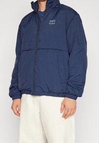 Navyblå vindjacka med dragkedja, elastiska ärmslut och en framficka. Tillverkad av lätt, strukturerat material med minimal logotyp.