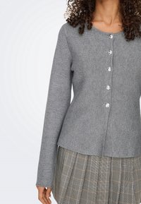 Femme portant un cardigan gris à manches longues boutonné et une jupe plissée à carreaux dans des tons gris, noir et beige.