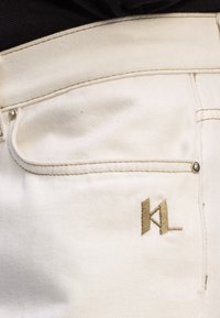 Suur lähivõte valge pükste taskust, millel on beežid õmblused ja beežist teibitud "KL" logo lähedal niidist.
