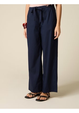 Femme portant un pantalon bleu marine à jambes larges avec une taille nouée, des sandales noires à brides et un bracelet rouge au poignet gauche, debout sur un sol beige.
