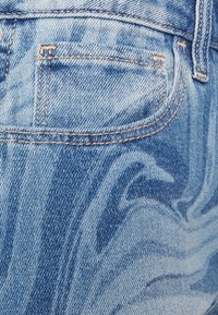 Calças de ganga azul desbotado com um padrão em espiral, apresentando um bolso traseiro e costura amarela em contraste. A textura parece suave e gasta.