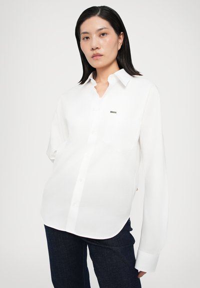 Valge pika varruka shirt nööpkaelusega, vasakul rinnal pehme tasku ja väike brändi silt. Sile tekstuur.