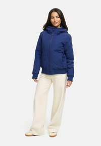 Marineblauwe ritsjack met een capuchon, ribbelmanchetten, zijzakken en een soepele textuur, gecombineerd met beige broek met wijde pijpen en lichte schoenen.