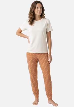 Vrouw staat blootsvoets, draagt een effen wit T-shirt met korte mouwen en roestkleurige joggingbroek met patroon tegen een witte achtergrond.