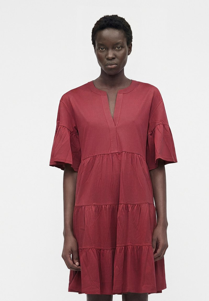 Γυναίκα που φοράει κόκκινο φόρεμα μέχρι το γόνατο, φαρδύ, με στρώσεις και μανίκια με βολάν μέχρι τους αγκώνες, στέκεται μπροστά σε απλό, φωτεινό φόντο.