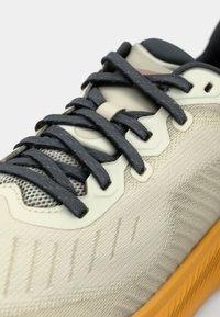 Hellbeige Sportschuh mit strukturiertem Obermaterial, grauen Schnürsenkeln und orangener Gummisohle. Verfügt über verstärkte Ösen und eine gepolsterte Ferse.