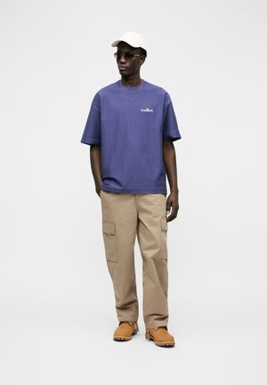 Uomo con cappellino bianco, occhiali da sole scuri, maglietta blu, pantaloni cargo beige, stivali marroni e catene d'argento, in piedi davanti a uno sfondo semplice.