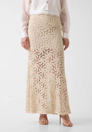 Donna che indossa una gonna maxi in pizzo beige lavorata a uncinetto, una camicetta bianca con bottoni e sandali beige con tacco alto, in piedi su uno sfondo bianco.