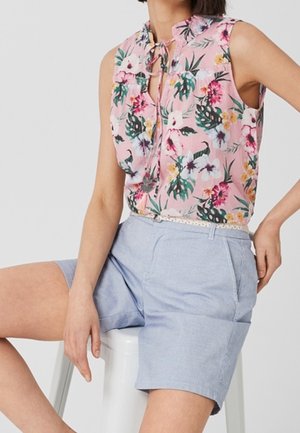 Mouwloze roze blouse met bloemenprint gecombineerd met lichtblauwe shorts met hoge taille, zittend op een witte kruk met één hand rustend op de dij.