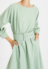 Miętowa sukienka o luźnym kroju, z szerokim dekoltem, długimi bufkami, wcięciem w talii i dopasowanym paskiem z kwadratową klamrą.
