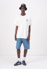 Balta medvilninė marškinėliai su juodu logotipu, šviesiai mėlyni denim šortai, juoda kibirėlio kepurė, balti kojinės, ir mėlyni Adidas sportbačiai.