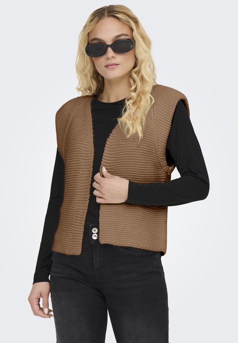 Bruine gebreide mouwloze vest met een gestructureerd patroon, gedragen over een zwart shirt met lange mouwen, gecombineerd met donkere spijkerbroeken en zwarte zonnebril.