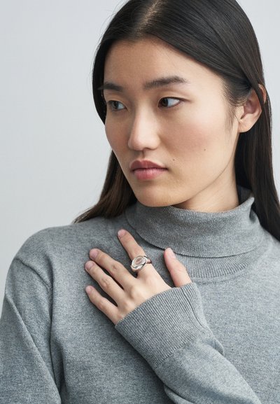 Ezüst gyűrű ovális, átlátszó kővel. A modellen szürke garbó pulóver van, keze a mellkasán, bemutatva az ékszert semleges háttér előtt.