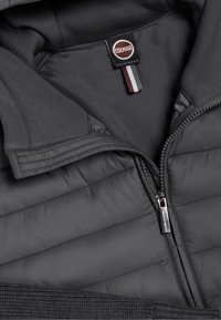 Dunkelgraue gesteppte Jacke mit teilweise geöffnetem Reißverschluss, gestrickten Bündchen und einem schwarzen Etikett mit der Aufschrift "Colmar Originals" am Krageninnenseite.