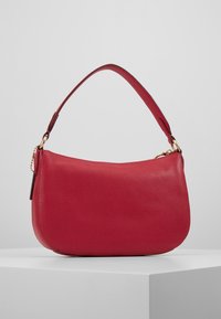 Sac à main en cuir rouge avec une forme doucement arrondie. Dispose d'une seule sangle large et d'accents métalliques dorés. Surface texturée.