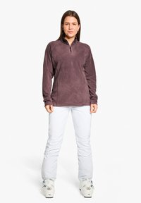 Lila Fleece-Pullover mit einem halben Reißverschlusskragen, kombiniert mit weißen Schneehosen und Skischuhen. Glatte Textur, leichtes Design.