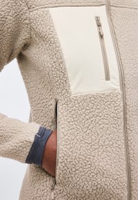 Άτομο που φοράει μπεζ υφασμάτινο μπουφάν από φλις με φερμουάρ στο στήθος και πλαϊνές τσέπες, τοποθετώντας το χέρι στην πλαϊνή τσέπη πάνω από ένα γκρι μανίκι.