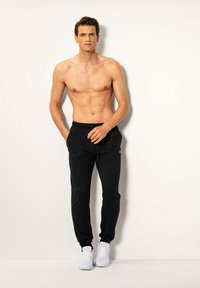 Schwarze Jogginghosen mit einem elastischen Bund, Seitentaschen und einem dezenten Logo. Getragen mit weißen Sport schuhen und einem nackten Oberkörper.